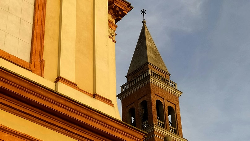 Una vista dal basso di una chiesa con una facciata gialla e una torre campanaria in mattoni rossi con una guglia.