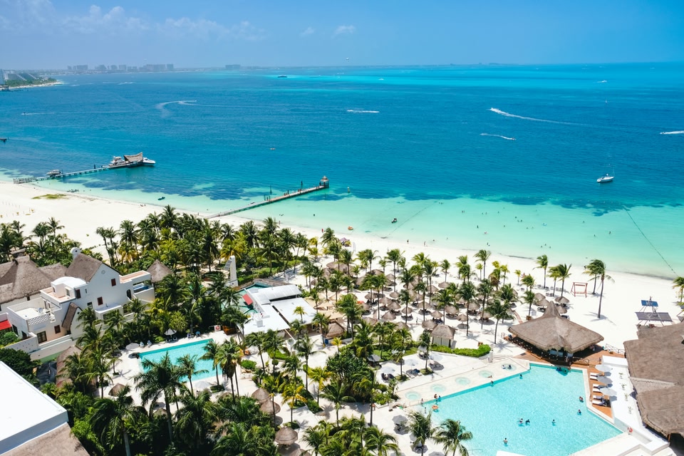 An aerial view of a tropical beachfront resort featuring white sand, turquoise water, swimming pools, and palm trees under a clear blue sky.
