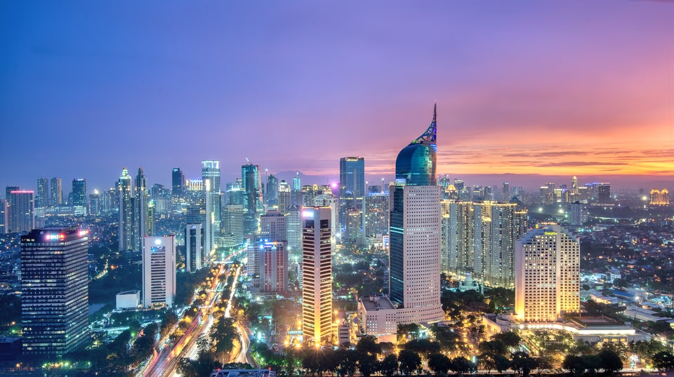 Skyline of Jakarta, Indonesia, at sunset with brightly lit skyscrapers, including the distinctive curved-top Wisma 46 building, and traffic trails below.