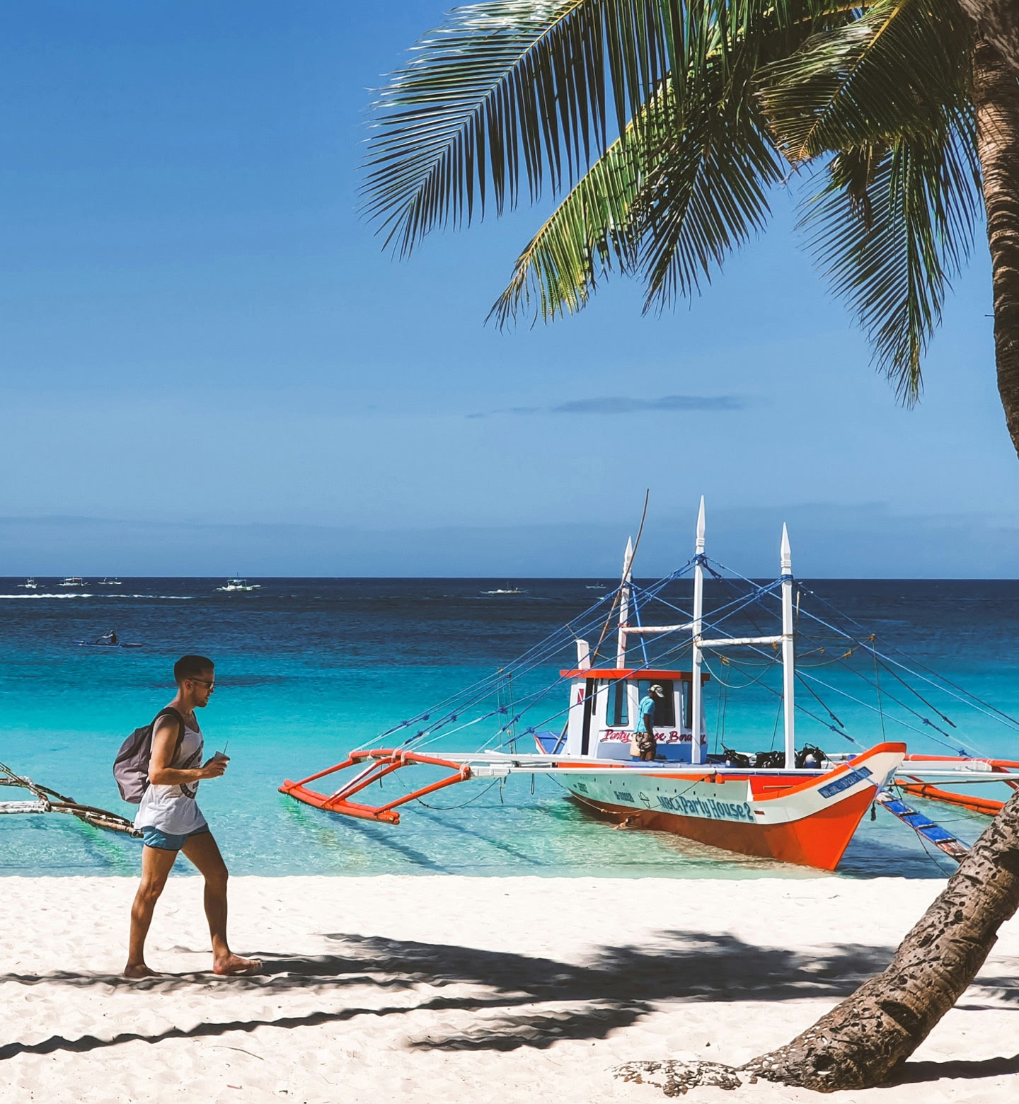 フィリピンのカティクランで、ヤシの木が茂る白い砂浜に係留されたボートの横を歩く男性。