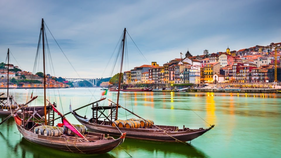 Colorful buildings of Porto reflected in the Douro River, with wine-carrying boats in the foreground.