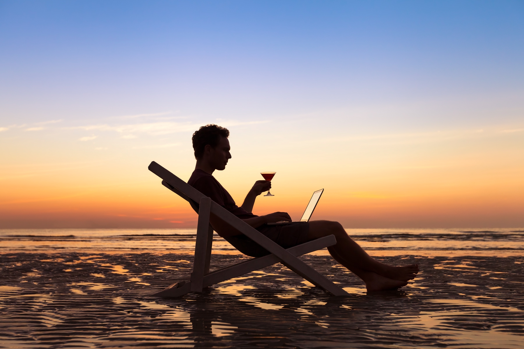 man sitting in lounge chair on beach working on a laptop during workcation