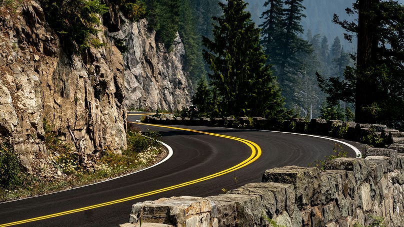 A scenic view of a winding asphalt road with yellow lines, bordered by a stone wall and a steep rock face. Lush green trees cover the surrounding mountains.