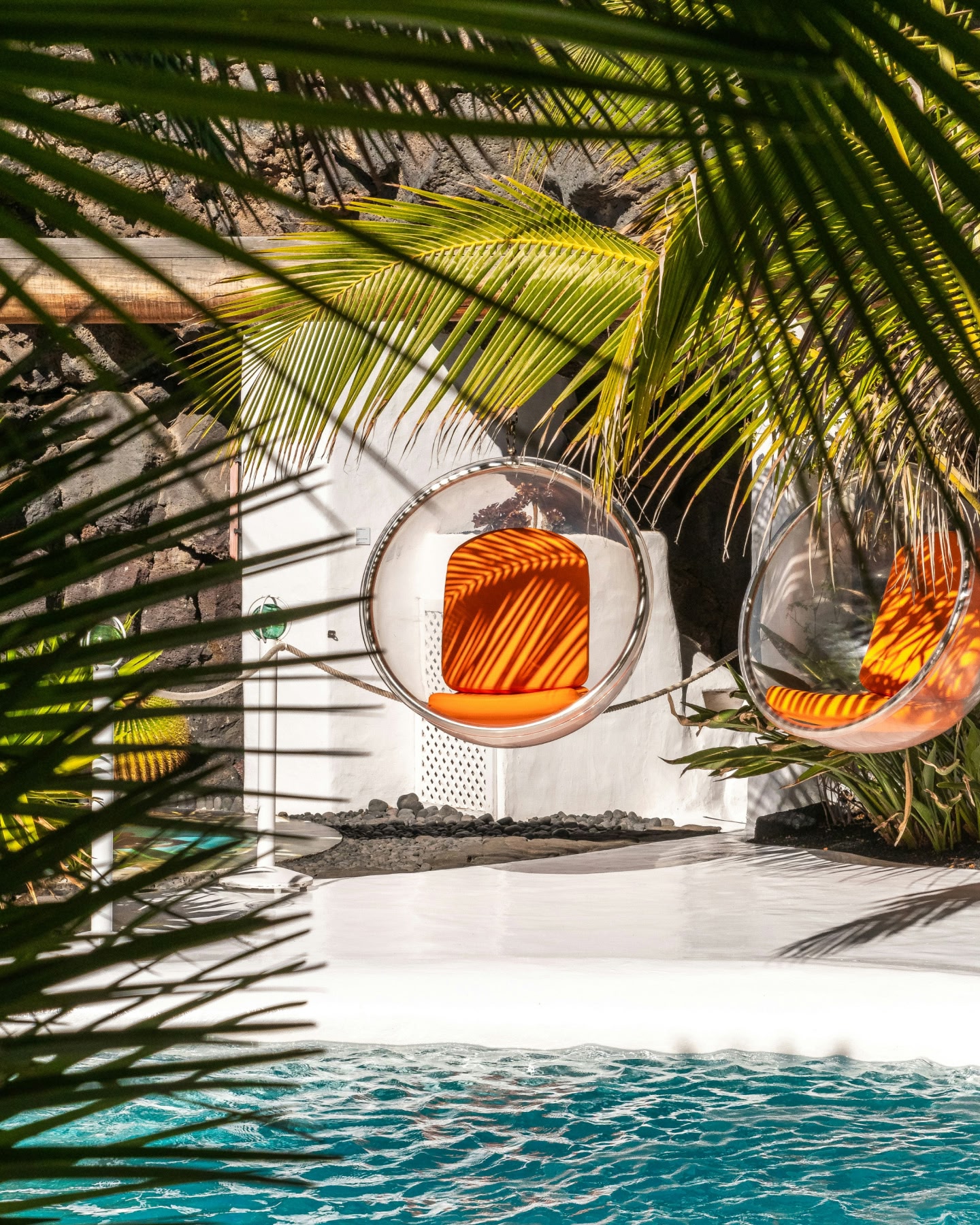 ヤシの木陰のプールの横にある、2つのオレンジ色のクッション付きハンギングチェア。