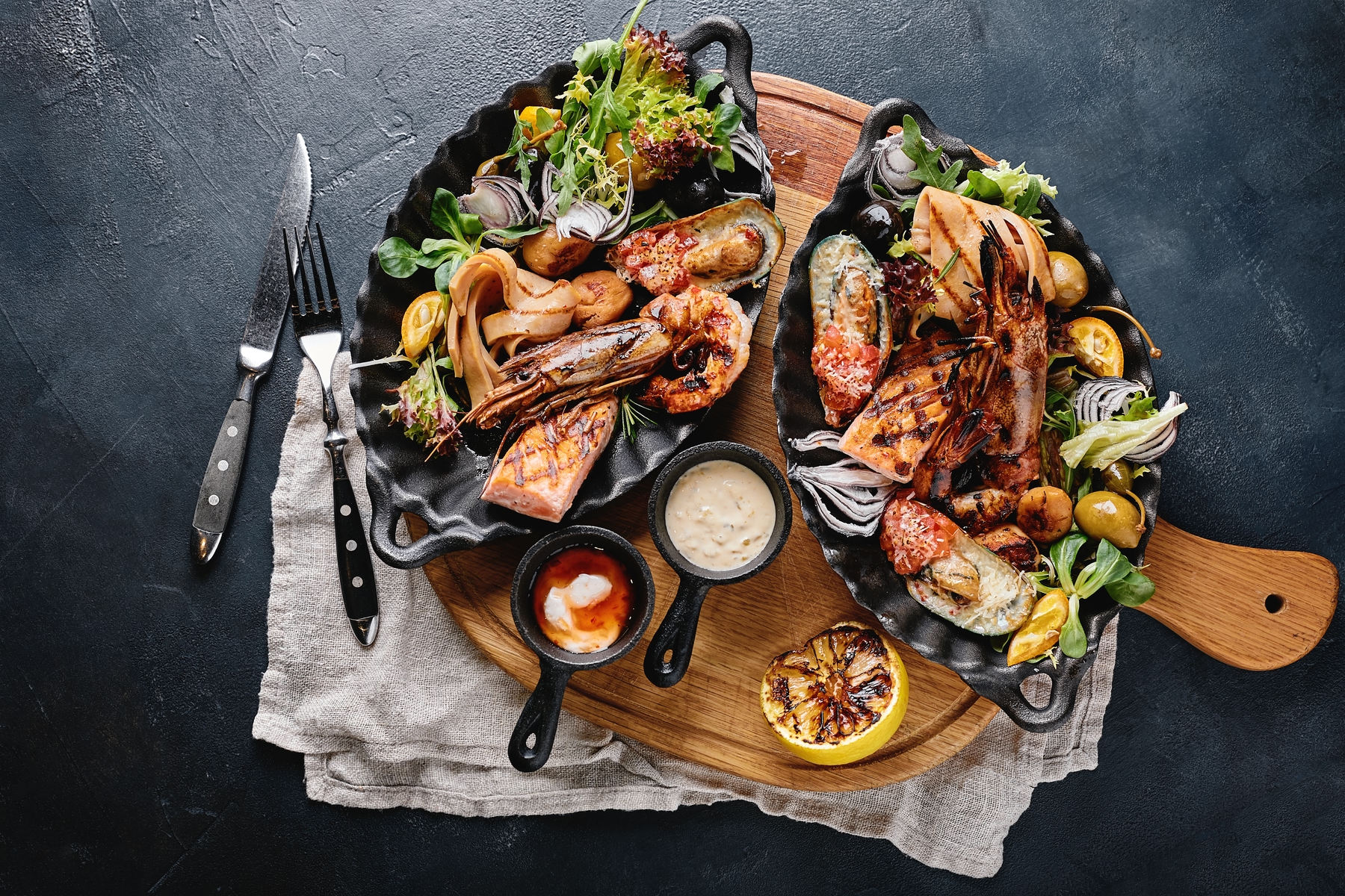 Slate table with wood board and dark-colored plates filled with grilled seafood and vegetables 