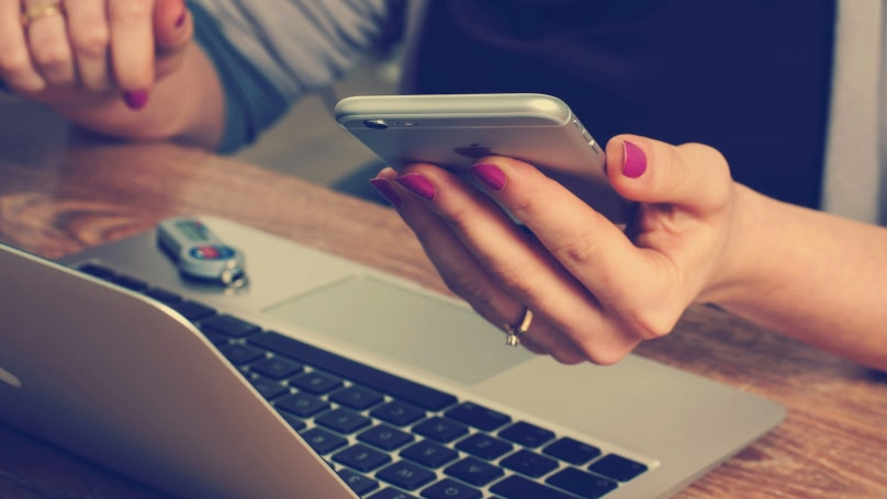 A woman with reddish-purple nail polish holds a smartphone while working on a silver laptop at a wooden desk.