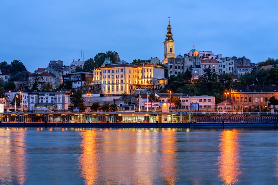 Evening view of Belgrade’s historic skyline with the illuminated Church of St. Michael and surrounding buildings reflecting on the Sava River.