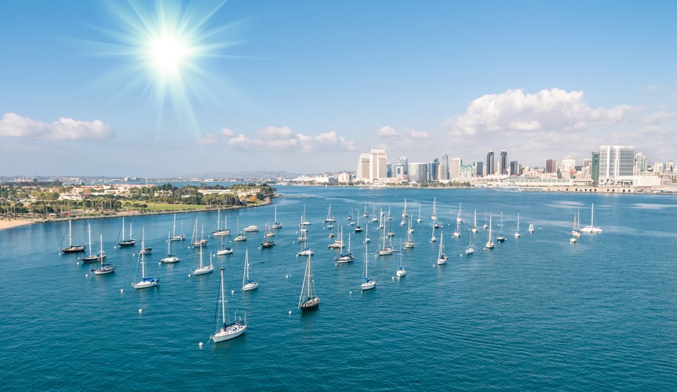 Aerial view of numerous sailboats anchored in a blue bay under a bright sun, with the San Diego city skyline in the background.