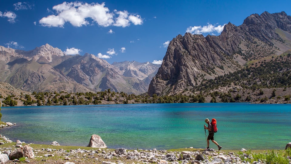A hiker walks along a turquoise lake with mountains in the background.