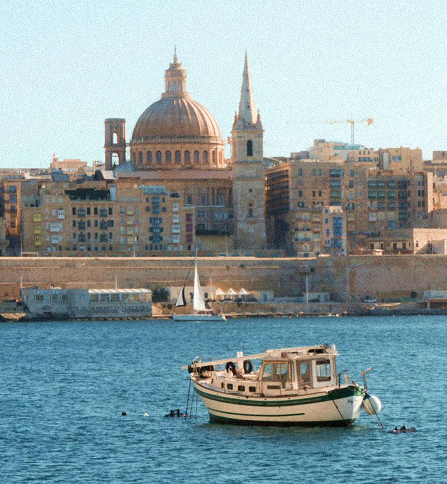 Łodzie unoszą się na błękitnych wodach, a w tle widać kopuły budynków w Valletcie na Malcie.