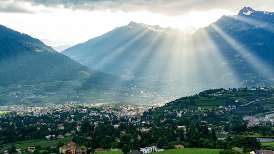 Paesaggio alpino con un paese in una valle e raggi di sole che filtrano tra le montagne.