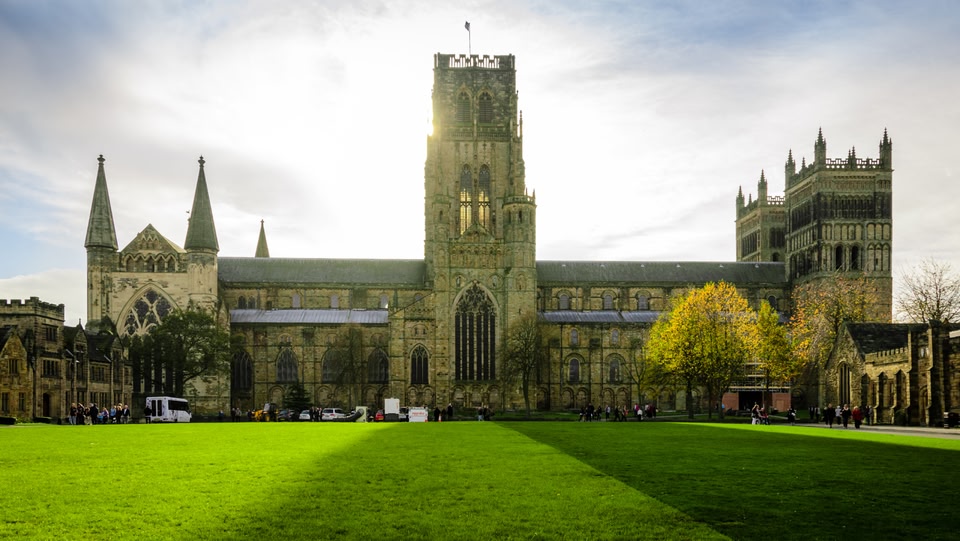 Durham Cathedral, a magnificent example of Norman architecture, dominates the landscape. Its imposing towers and intricate stonework are visible against a backdrop of a bright sky and a green lawn.