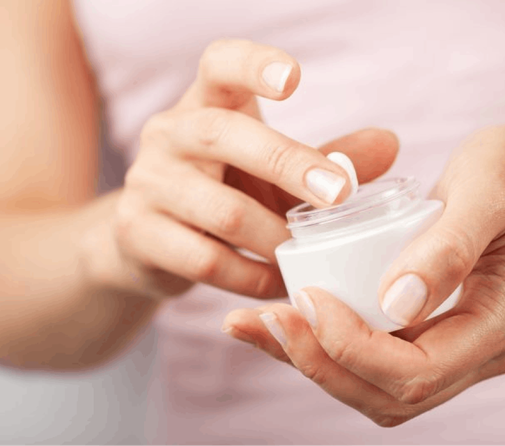 A close-up of a woman's hands delicately holding a pot of cream, as she scoops a bit out with her finger.