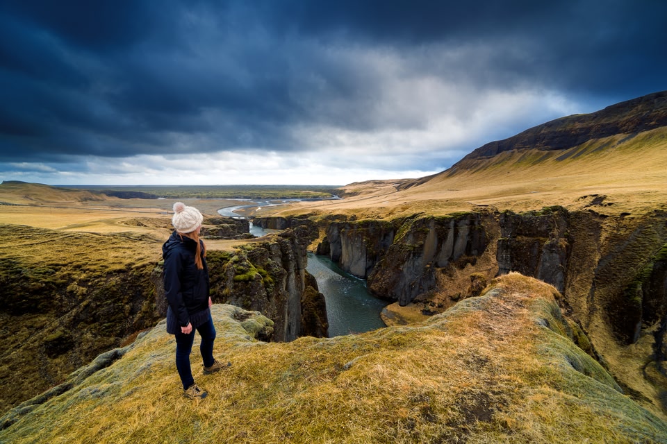 Young woman wearing black coat and white hat looks out on dramatic landscape of Fjadrargljufur canyon under dramatic stormy skies.
