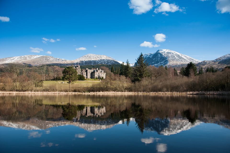 Inverlochy Castle, with snow-capped mountains in the background and a reflectieve loch in the foreground.