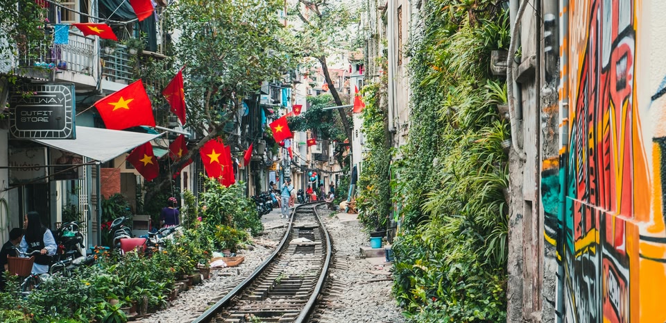 Vía de tren estrecha que atraviesa una calle residencial, rodeada de banderas vietnamitas, abundante vegetación y muros con grafitis coloridos.