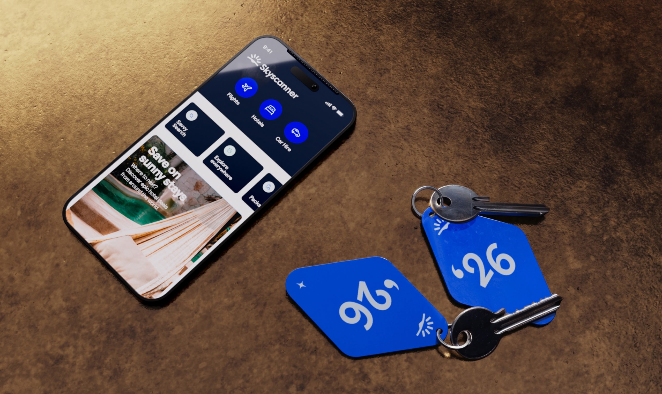 スカイスキャナーのロゴ入りキーホルダーが付いたホテルの客室の2つの鍵の横にある、スカイスキャナーアプリが表示されたスマートフォン。