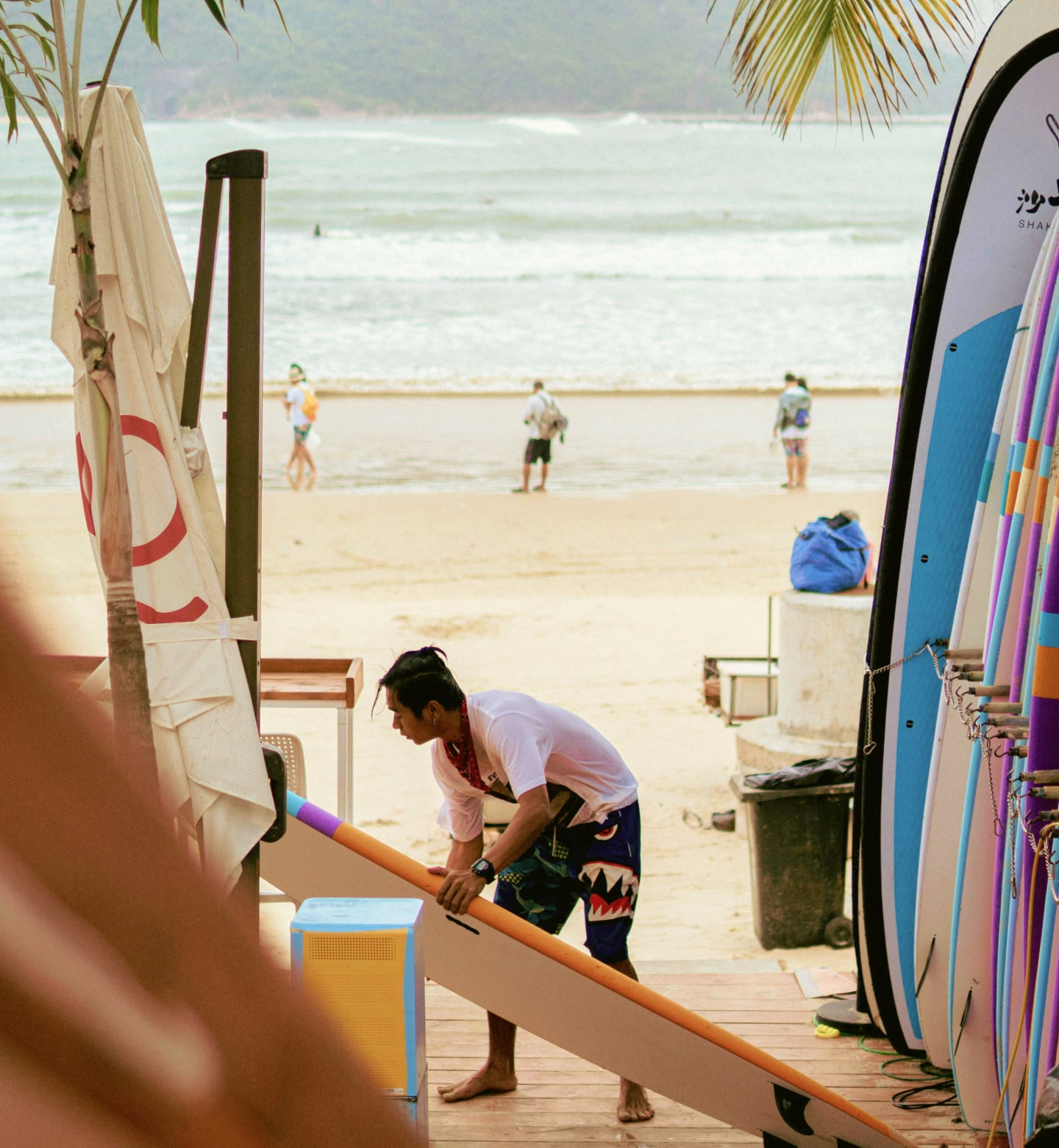 Un homme à Sanya, en Chine, prépare une planche de surf sur la plage tandis que d'autres marchent le long du rivage.