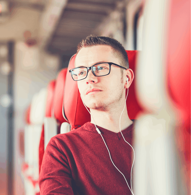 A man wearing a red t-shirt and glasses relaxing on a train listening to music on some headphones.