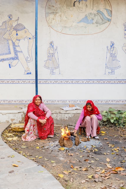 Two women wearing bright red and pink sarees seated around a small open fire in a courtyard at Dera Mandawa, Jaipur, with traditional blue-and-white Rajasthani murals of elephants and figures painted on the walls behind them.