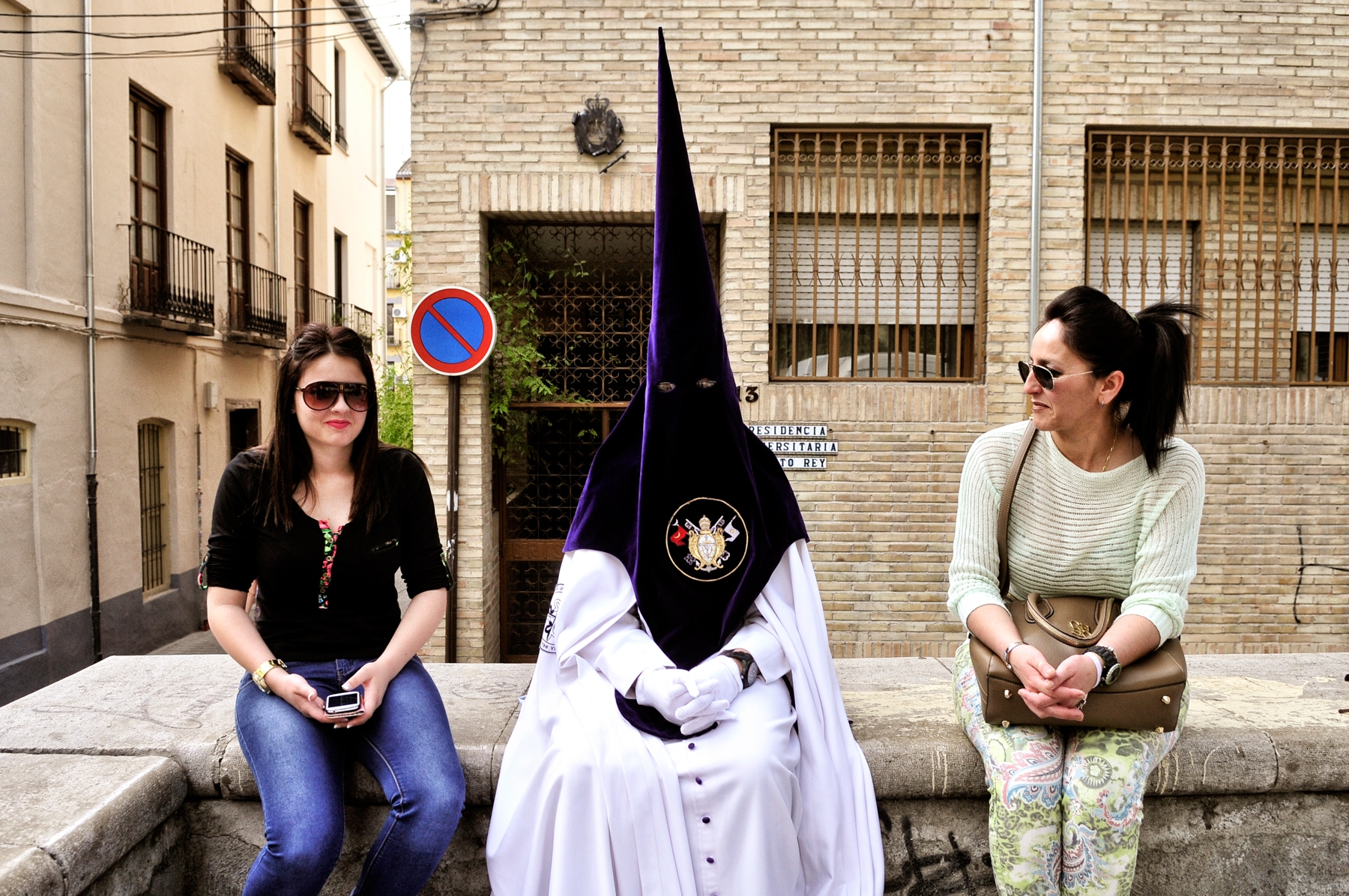Un nazareno espera el inicio de su procesión en la calle ante la atenta mirada de dos turistas © Bisual Studio