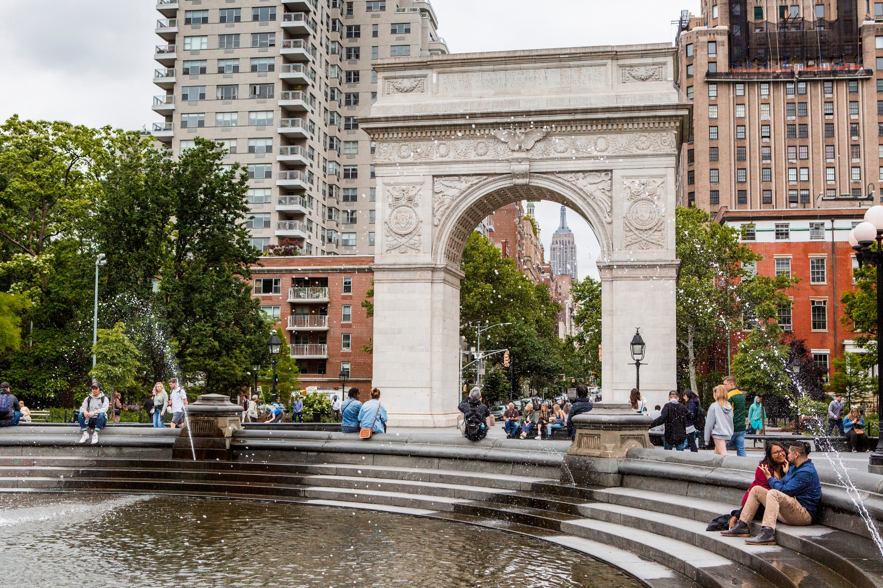 View of the arch and fountain in Washington Square park with people sitting on the steps and walking behind