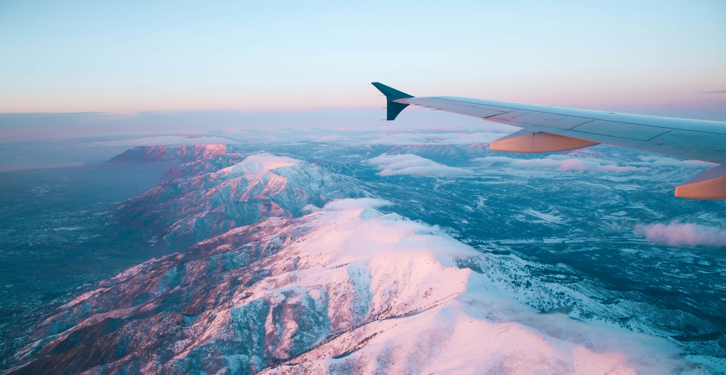 Birds eye view of snowy mountain range. An aeroplane wing is seen at the corner of the image. 