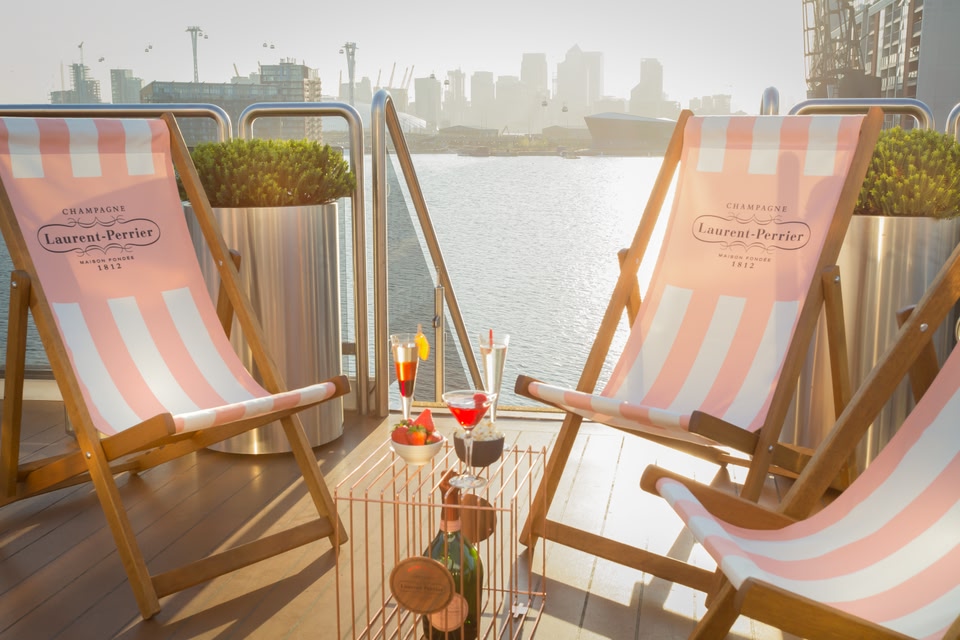 Pink and white stripe Laurent Perrier deckchairs on the Sunborn London Yacht Hotel, with cocktails on the table in front and the London city skyline behind.