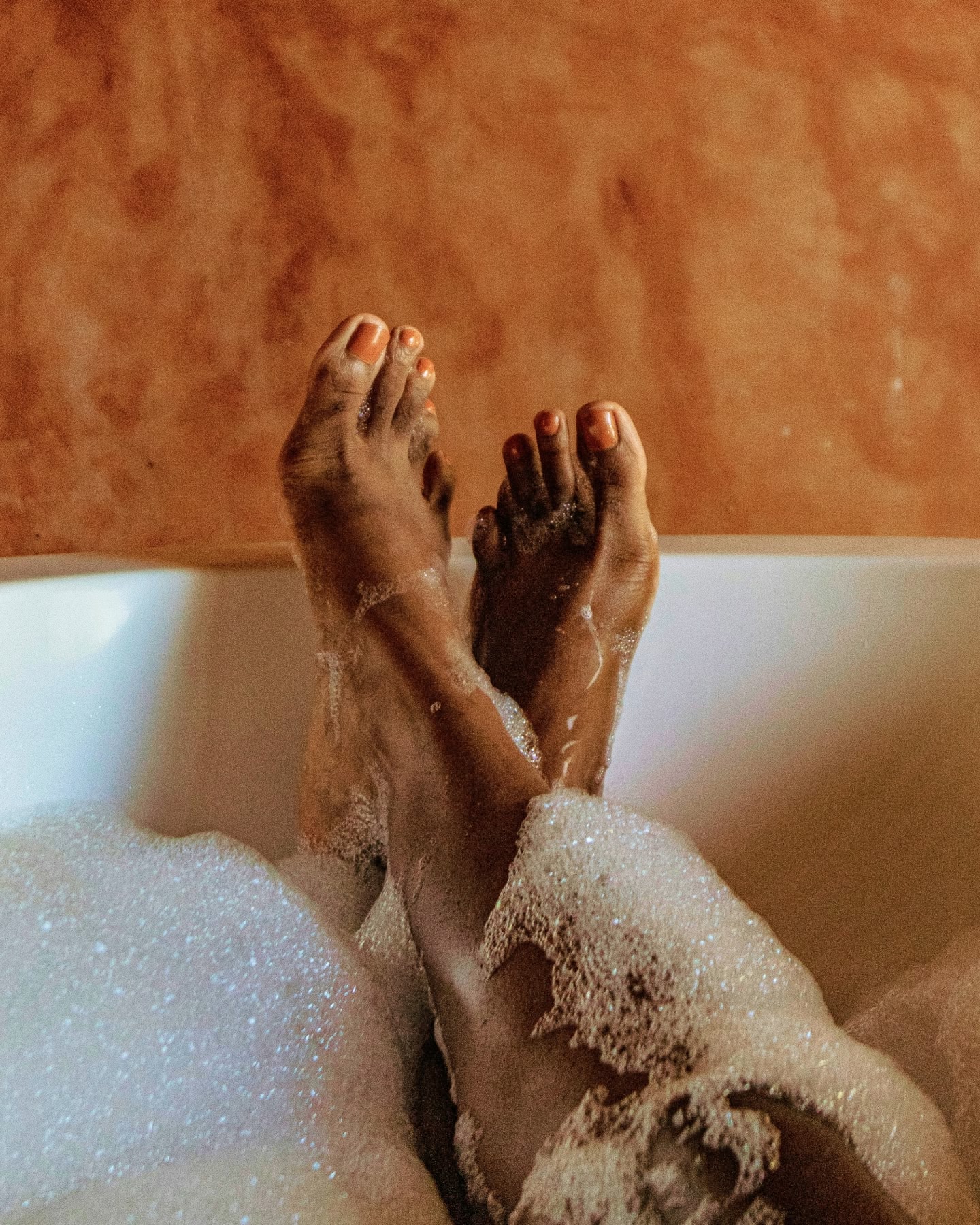 Los pies y las piernas de una mujer cubiertos de burbujas, apoyados en el borde de una tina blanca junto a una pared naranja