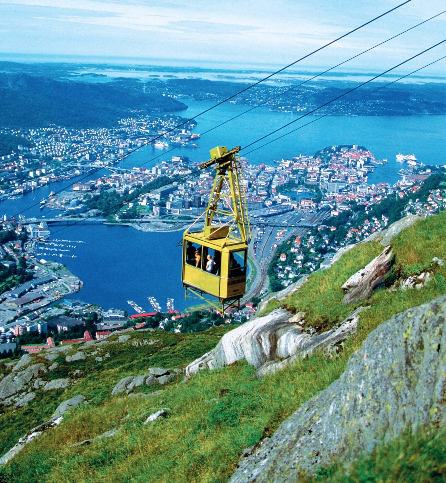 A yellow cable car suspended above a green, rocky mountain, overlooking the city of Bergen, Norway