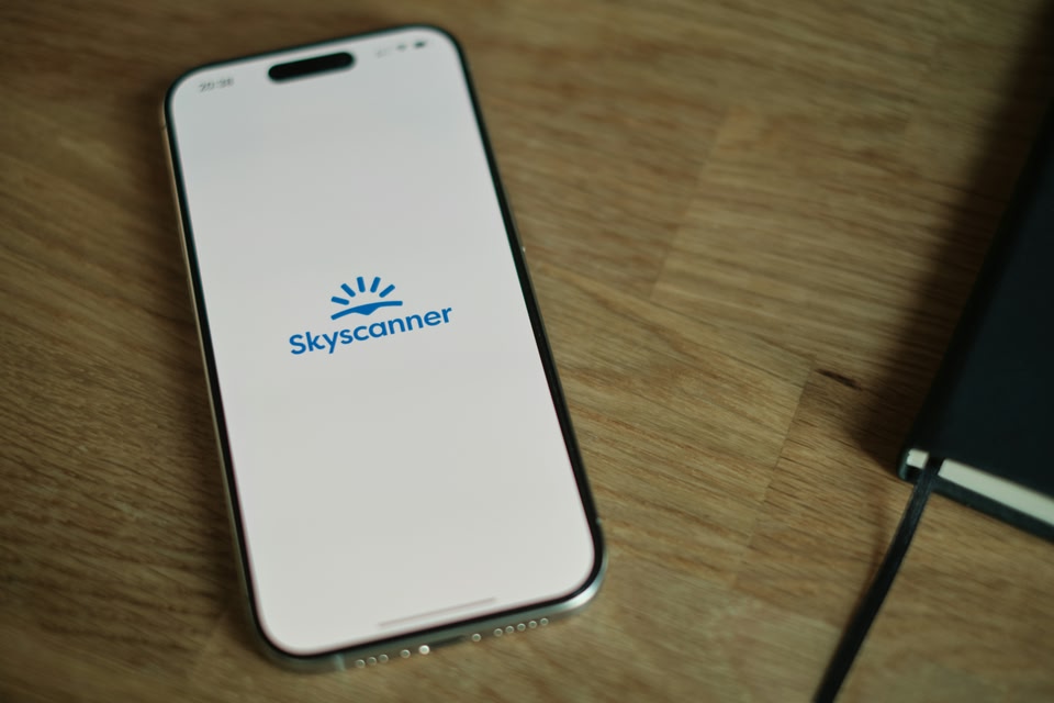 An iPhone displaying the Skyscanner app logo rests on a wooden surface next to a black notebook.