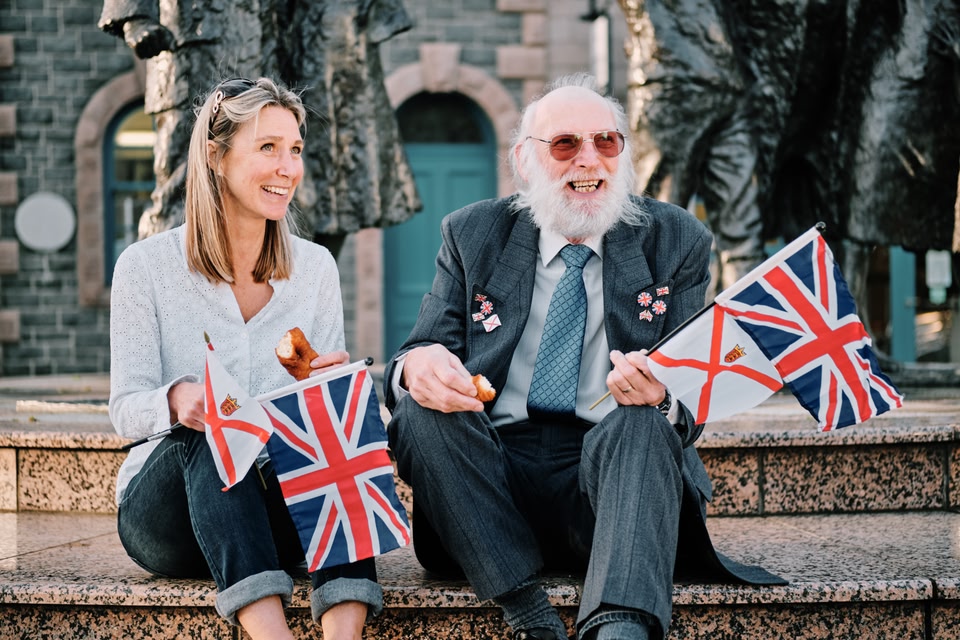 Young woman and older man sitting on steps, holding the Jersey flag and Union Jack.