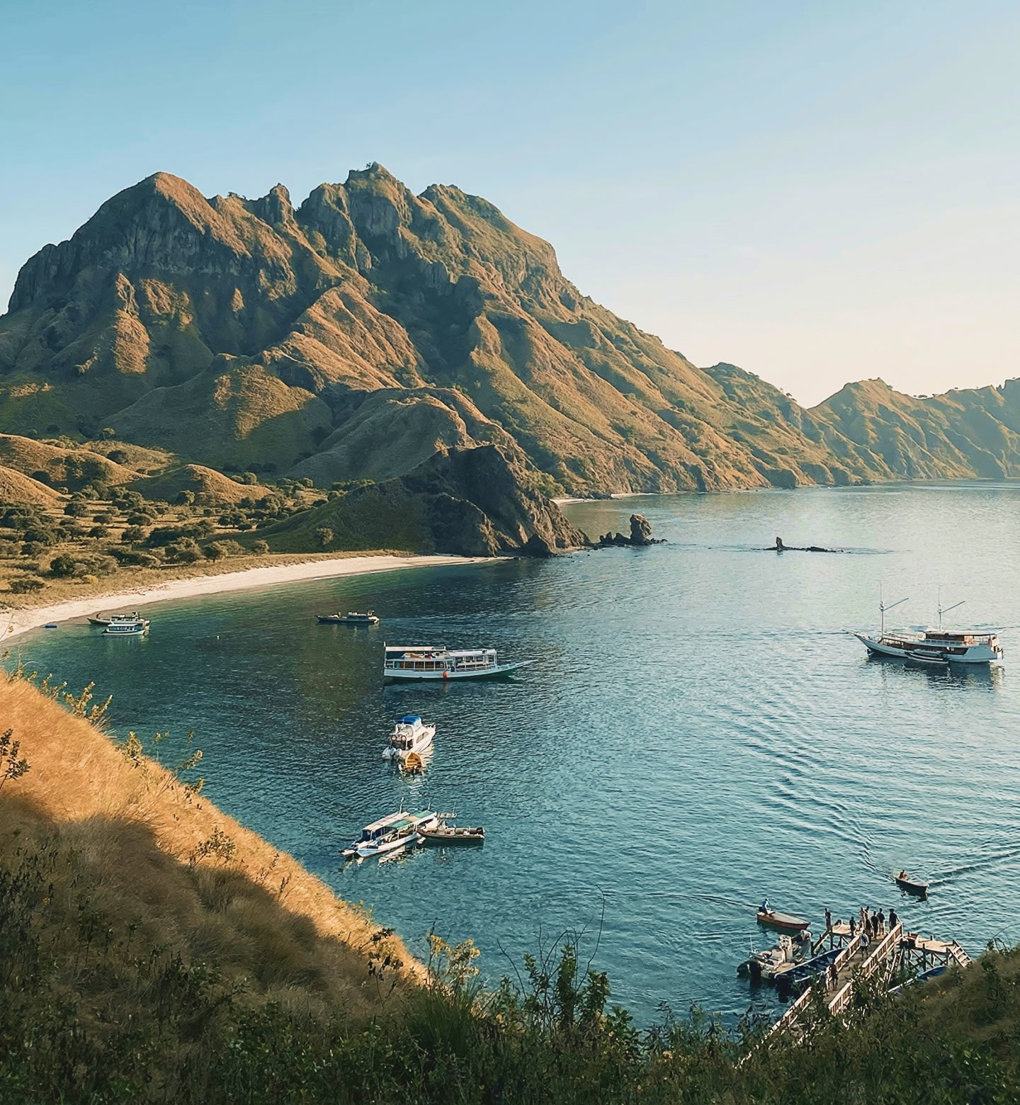 A white sandy beach in a bay with small boats floating on the ocean, with a mountain in the background in Padar Island in Labuan Bajo, Indonesia