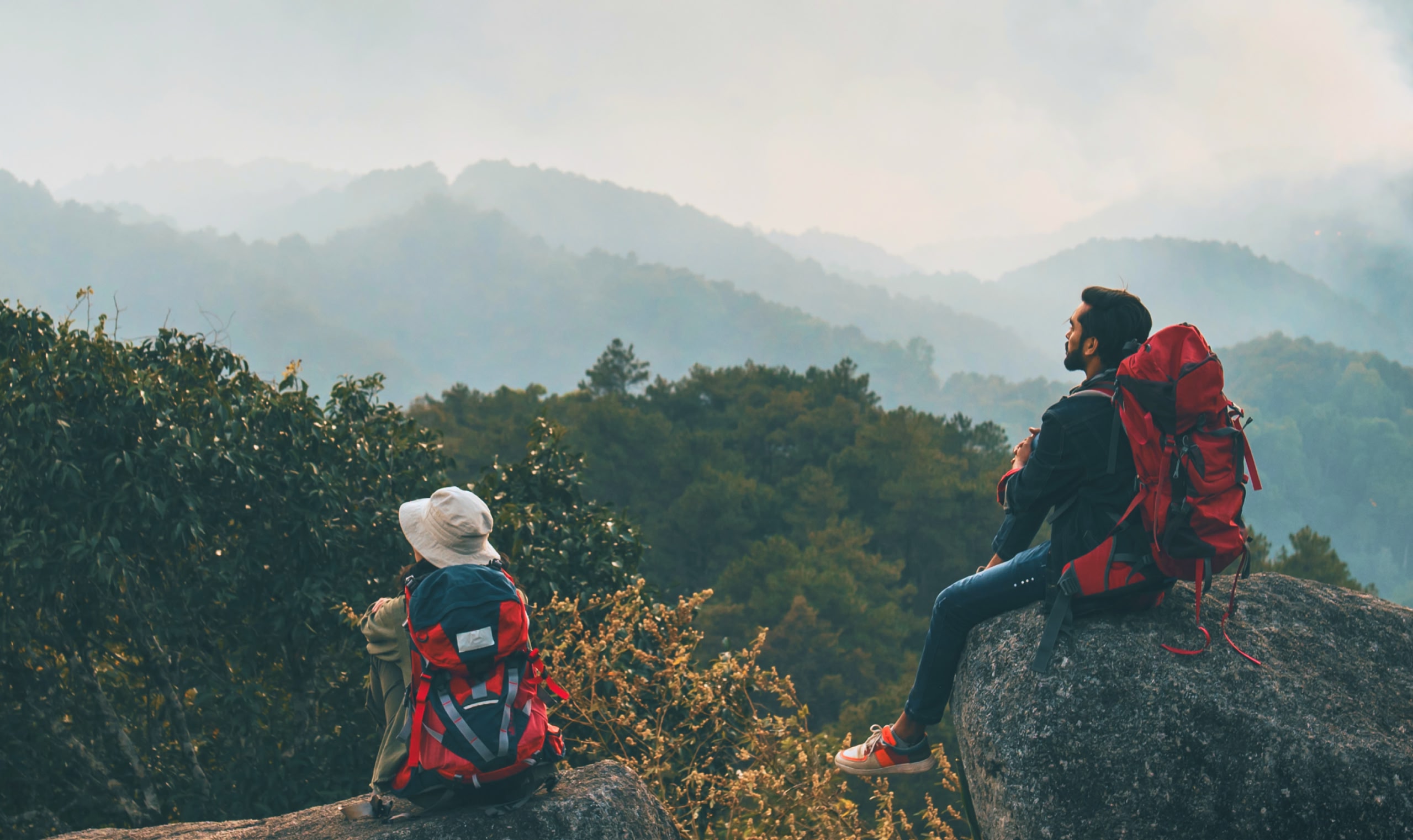 A man wearing a red backpack sits on top of a rock, looking out over trees and hills covered in light mist.
