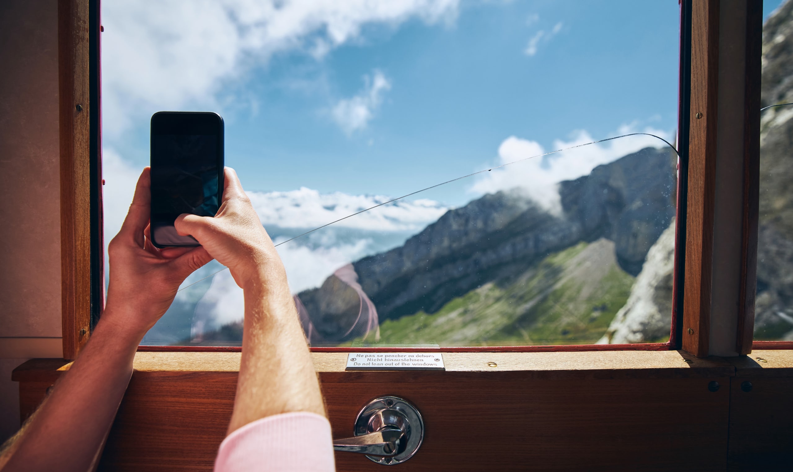 窓から山と空の景色をスマートフォンで撮影している手。