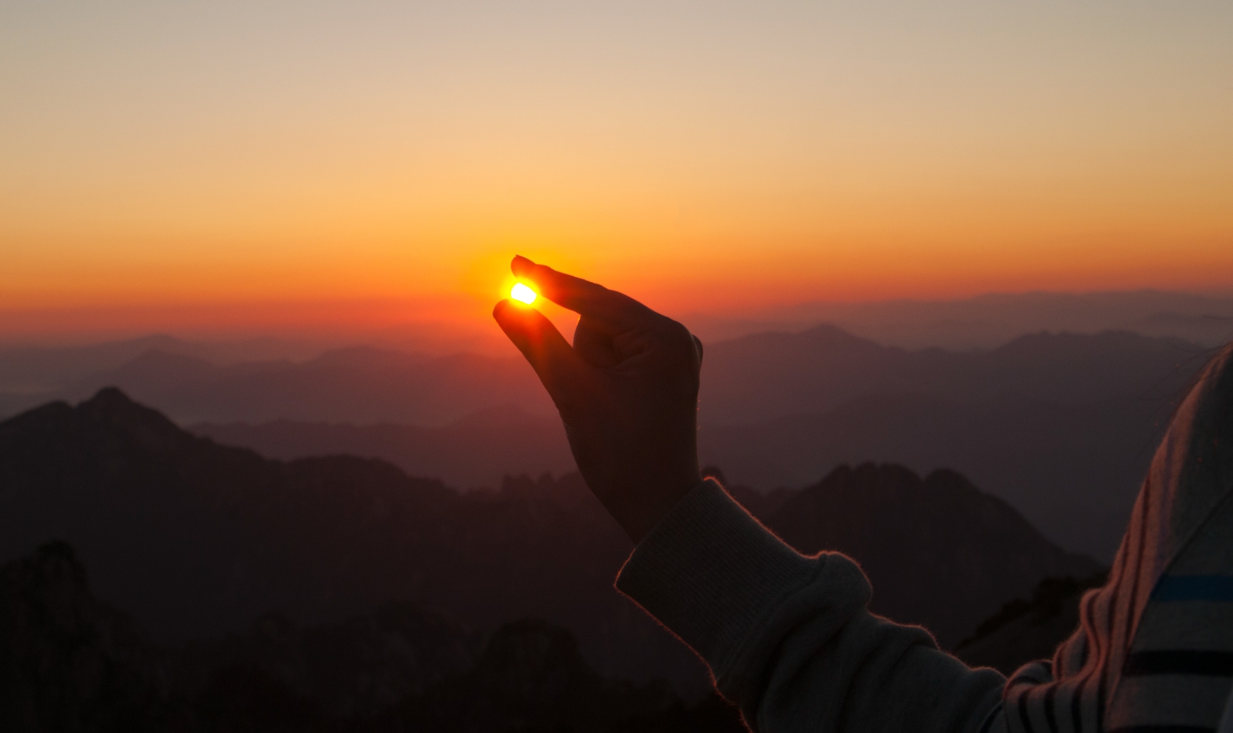 A hand grabbing at the setting sun over mountains.