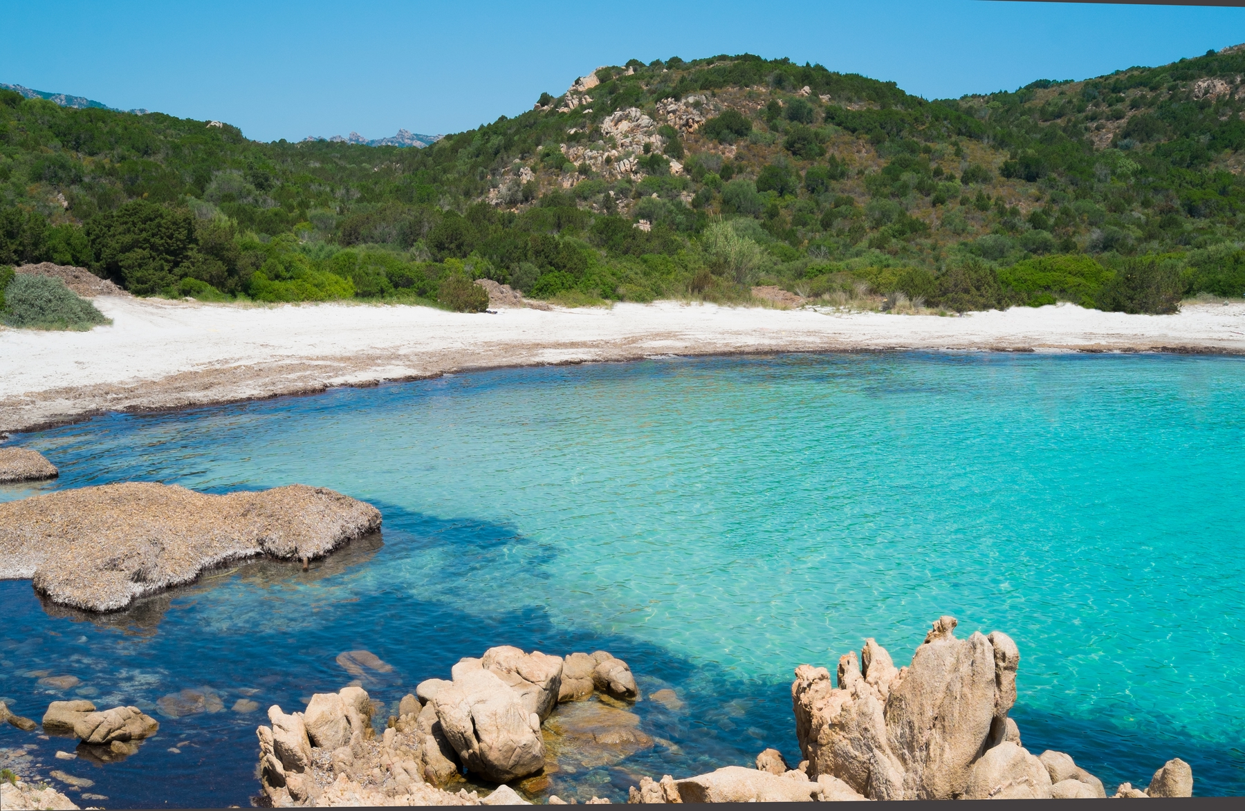 Picture shows a beach in Sao Tome and Principe, with bright blue waters, natural rocks, golden sands, and a backdrop of lush green hills. The sun is shining.