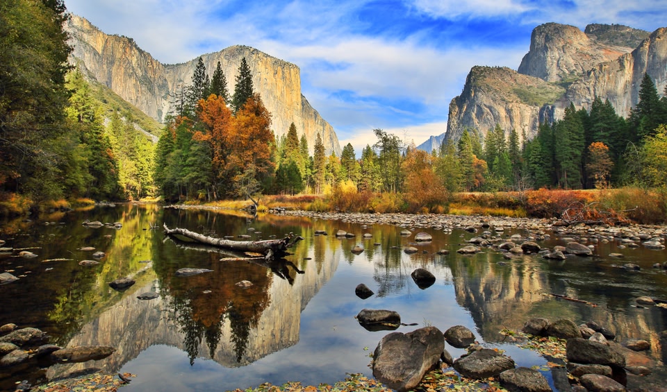 The towering granite cliffs of Yosemite Valley are reflected in the calm waters of a river, surrounded by vibrant autumn trees and a clear blue sky.