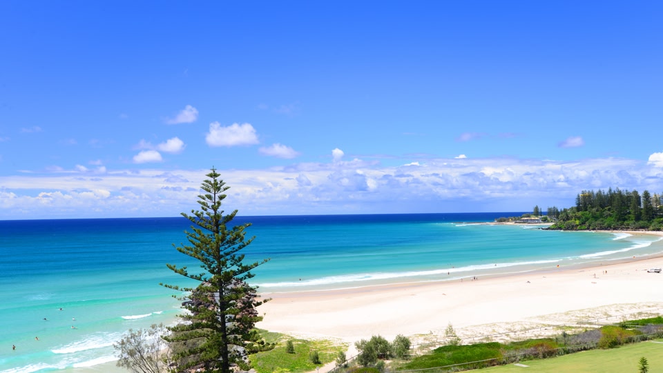 An image of a beach with blue water, white sand and green trees around it
