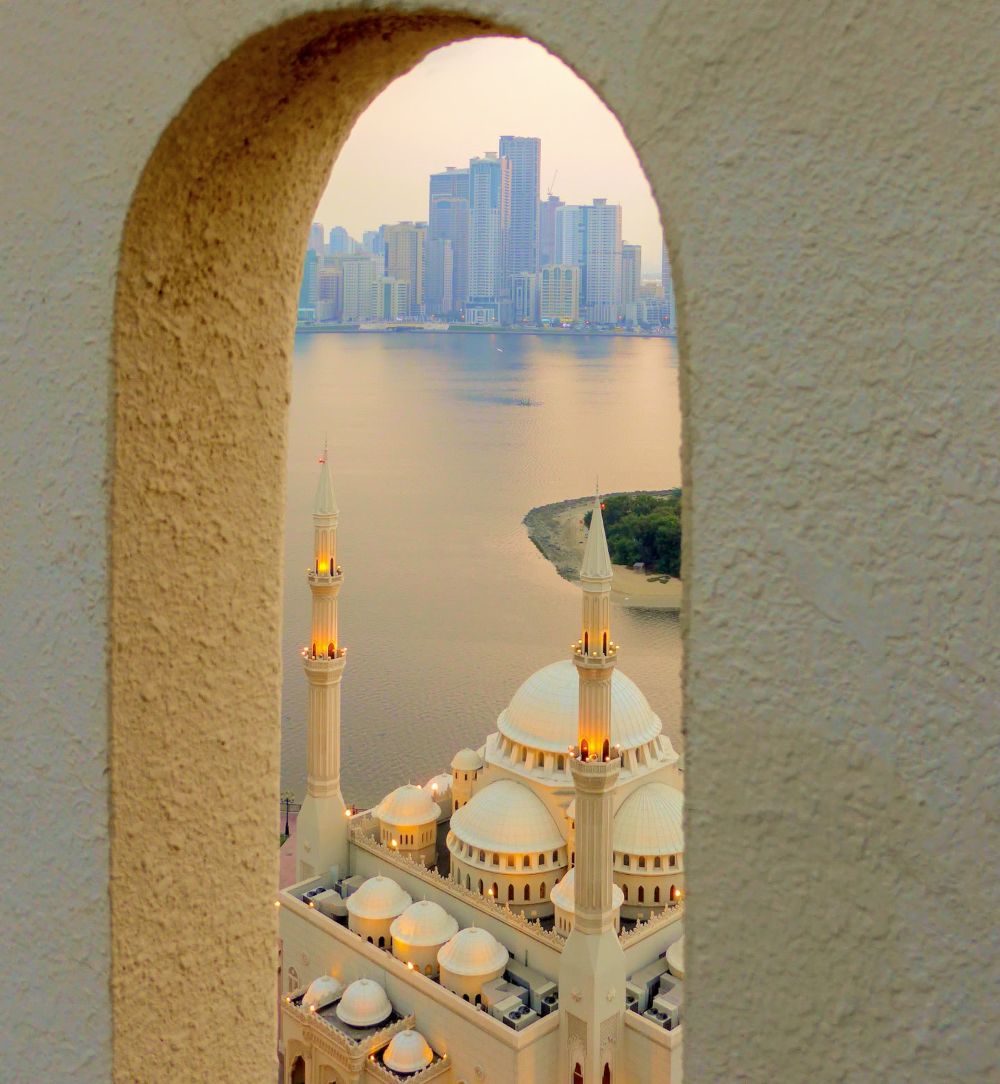 Weiße Moschee in Schardscha, Vereinigte Arabische Emirate, eingerahmt von einem Torbogen, mit Skyline der Stadt und Wasser dahinter.