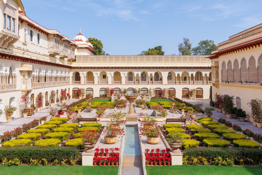The beautifully manicured formal garden courtyard of Rambagh Palace in Jaipur, viewed from an upper level, with symmetrical flower beds, a long central water channel, and the palace's white arched colonnades framing the space.