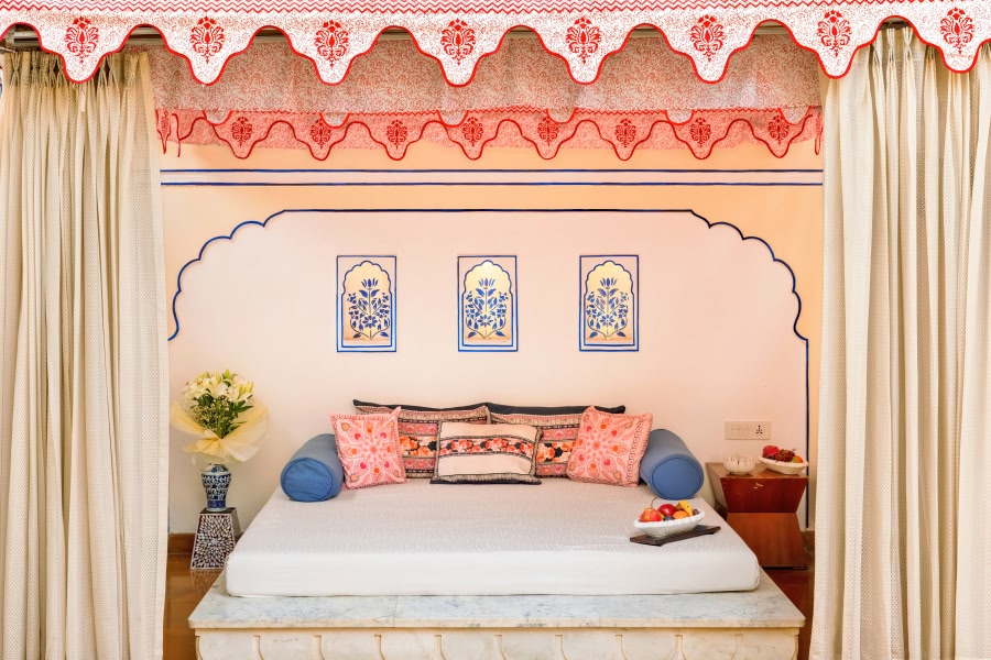 A luxurious hotel bedroom at The Leela Palace in Jaipur, styled with a canopied bed, colorful embroidered cushions in pink and blue, hand-painted blue floral wall panels, and a decorative Mughal arched headboard alcove.