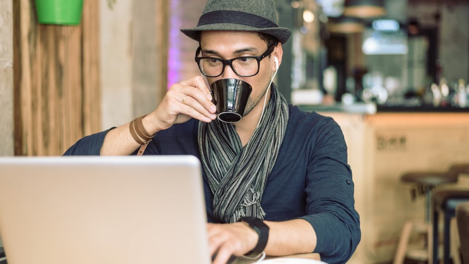 Person wearing glasses and a hat sipping a mug looking at a laptop screen
