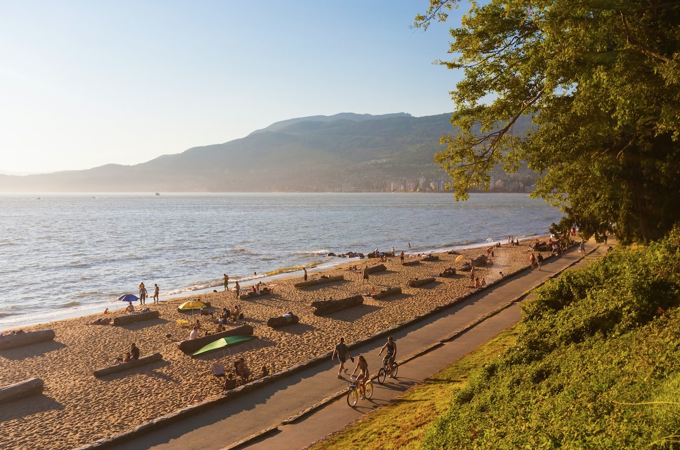 aerial view of Third Beach in Vancouver, driftwood logs on the ground and people biking down the Stanley Park Seawall, mid afternoon sun.