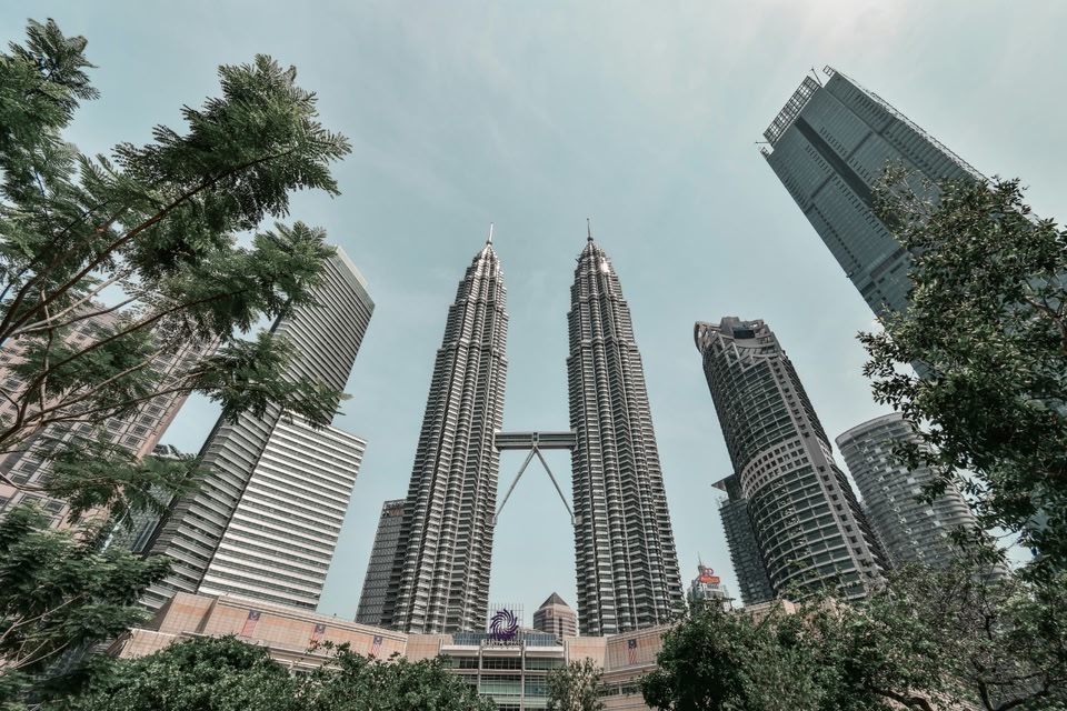A low-angle view of Kuala Lumpur's Petronas Twin Towers connected by a skybridge, surrounded by modern skyscrapers and lush green trees against a clear sky.