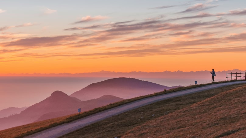 Una vista panoramica di montagne al tramonto, con una persona in piedi su una strada in primo piano. Il cielo è colorato di arancione e rosa, e le montagne sono avvolte nella foschia.