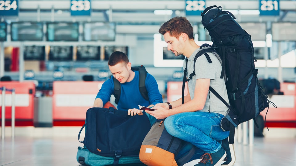 Deux voyageurs avec des sacs à dos sont agenouillés dans un terminal d'aéroport, en train de vérifier leurs passeports.