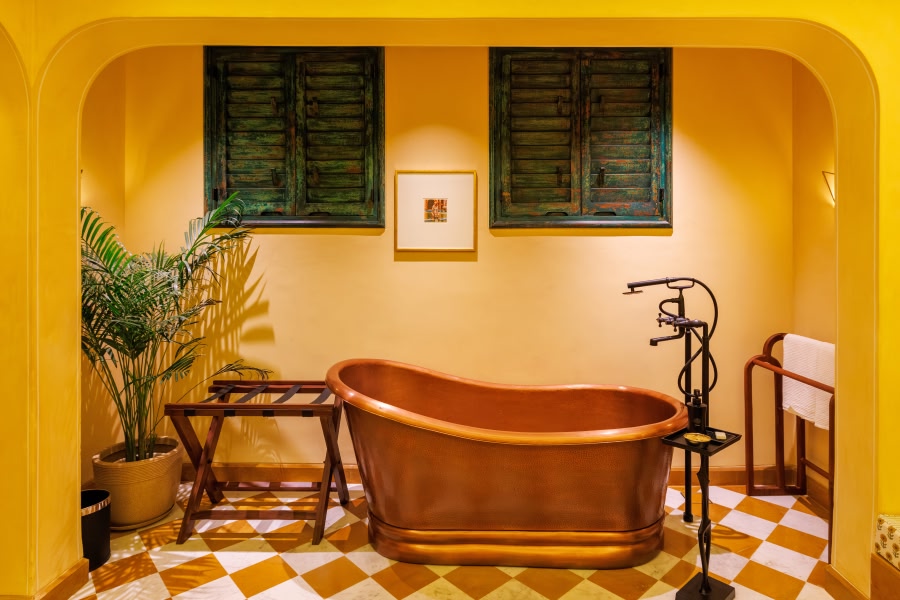 A gleaming copper freestanding soaking tub in a boutique hotel bathroom at Abode Jaipur, with bold yellow walls, a checkered terracotta and white tile floor, green-shuttered windows, and a lush potted palm plant.
