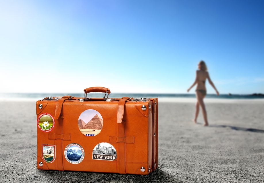 Suitcase with stickers on is sitting on a beach, as a girl is walking towards the sea