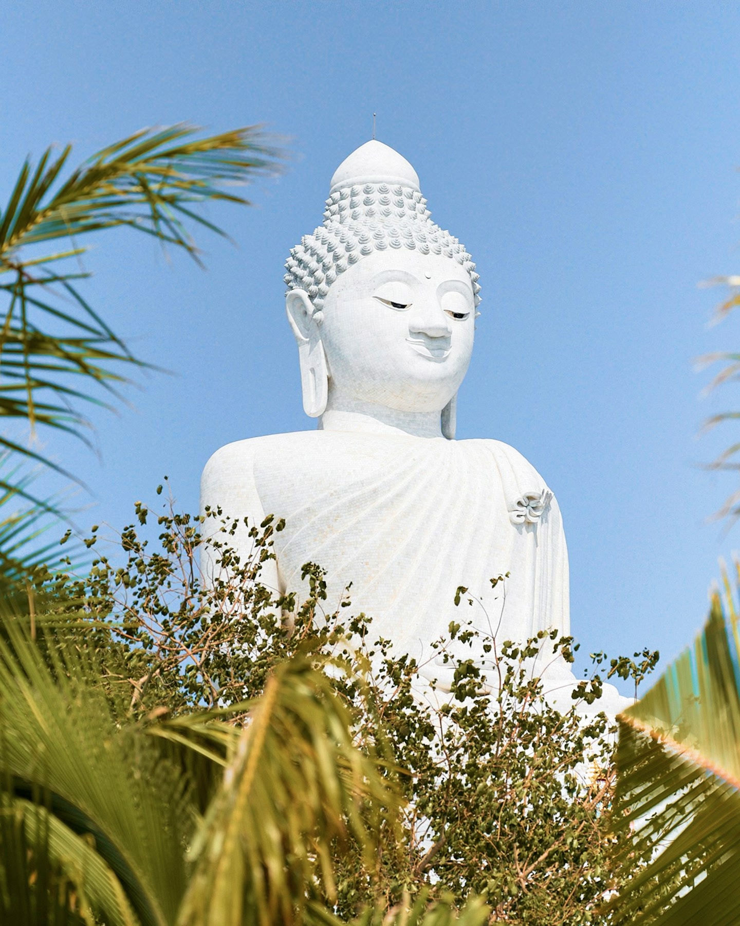 White marble Buddha peeks through palm trees under a bright sky in Phuket, Thailand.